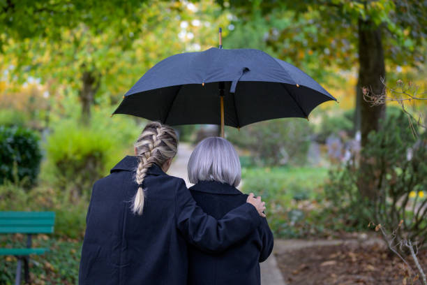 Close up rear view of daughter holding her mother on the way to the man's grave Close up rear view of daughter holding her mother in her arms on the way to the man's grave in the cemetery on a cloudy cold day annoying mother in law stock pictures, royalty-free photos & images