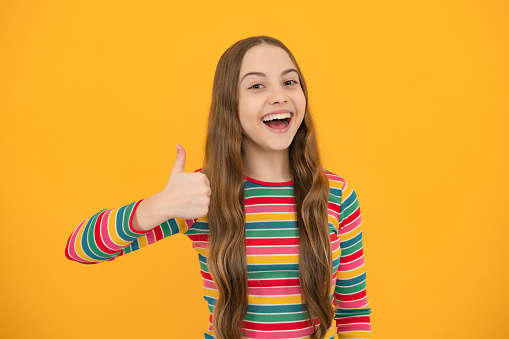 Teenager child girl 12, 13, 14 year old doing thumbs up gesture. Cheerful teenager girl showing thumbs up sign, isolated on yellow background