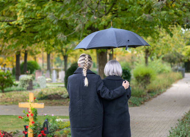 Rear view of daughter holding her mother on the way to the man's grave Rear view of daughter holding her mother in her arms on the way to the man's grave in the cemetery on a cloudy cold day annoying mother in law stock pictures, royalty-free photos & images