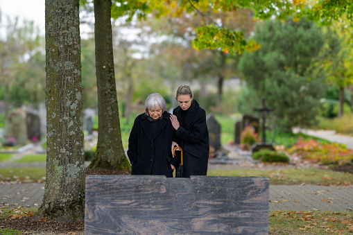Mother and daughter standing at the man's grave in the cemetery on a cloudy cold day and looking down