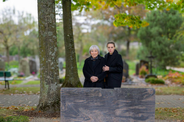 Mother and daughter standing at the man's grave and looking to camera Mother and daughter standing at the man's grave in the cemetery on a cloudy cold day and looking to camera annoying mother in law stock pictures, royalty-free photos & images