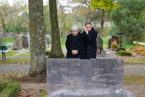 Mother and daughter standing at the man's grave in the cemetery on a cloudy cold day, the daughter wiping a tear from her face