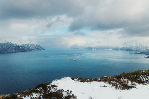 High-angle photo of the ferry boat sailing through dramatic winter fjord with crystal blue ocean water and view of snowcapped mountains during scenic sunset in More og Romsdal, Scandinavia