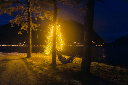Silhouette of female feeling happy lying in the camping hammock by the fjord near the lighting Chrismtas pine tree at night in Norway, Scandinavia