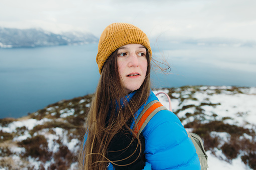 Portrait of female in yellow hat and blue winter jacket feeling freedom getting to the top of the snowy mountain peak admiring scenic view of the stormy ocean and islands in Norway, Scandinavia