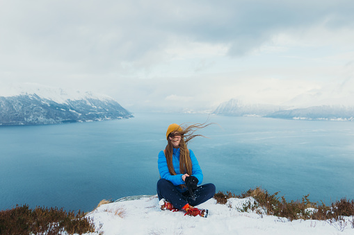 Front View of smiling female in yellow hat and blue winter jacket feeling freedom sitting on the top of the snowy mountain peak admiring scenic view of the stormy ocean and islands in Norway, Scandinavia