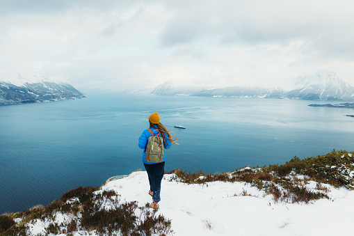 Rear View of female in yellow hat and blue winter jacket feeling freedom getting to the top of the snowy mountain peak admiring scenic view of the stormy ocean and islands in Norway, Scandinavia