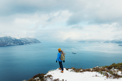 Side View of female in yellow hat and blue winter jacket feeling freedom getting to the top of the snowy mountain peak admiring scenic view of the stormy ocean and islands in Norway, Scandinavia