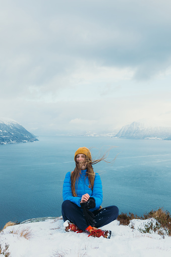 Front View of smiling female in yellow hat and blue winter jacket feeling freedom sitting on the top of the snowy mountain peak admiring scenic view of the stormy ocean and islands in Norway, Scandinavia