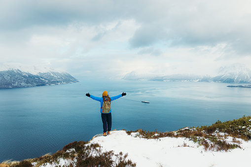 Rear View of female in yellow hat and blue winter jacket feeling freedom getting to the top of the snowy mountain peak admiring scenic view of the stormy ocean and islands in Norway, Scandinavia