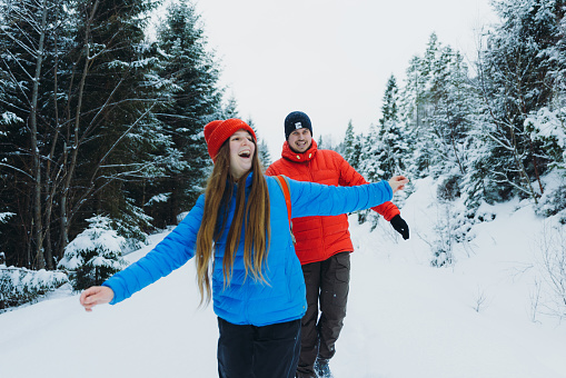 Front view of smiling female and male in colourful jackets contemplating the Christmas time outdoors having fun together and walking through the deep snow in mountain pine woodland in Norway, Scandinavia