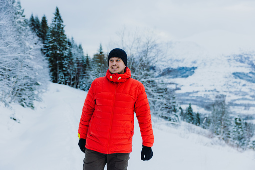 Front View of a smiling man in red jacket contemplating the Christmas time outdoors walking in the deep snow in pine woodland with Mountain View of Norway, Scandinavia