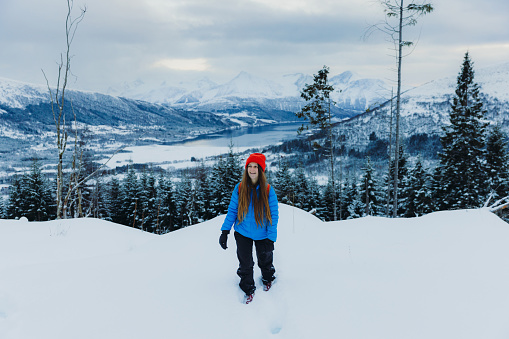 Front view of smiling female in red hat and blue jacket enjoying the Christmas time outdoors hiking in the pine forest contemplating the view of the snowcapped mountains and frozen reflection lake woodland above in Norway, Scandinavia