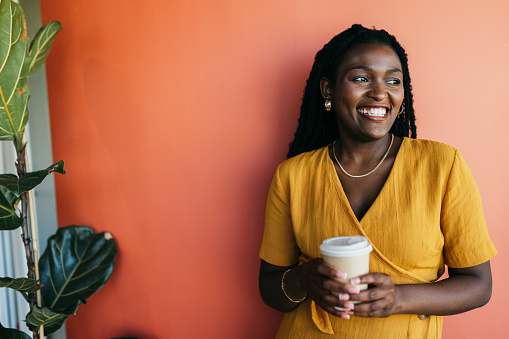 Cheerful young woman with afro hair holding coffee cup, smiling and looking sideways against orange background