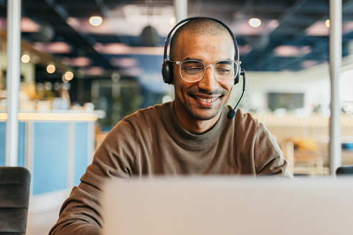 Businessman wearing eyeglasses and headset, smiling cheerfully and working with laptop in call center office