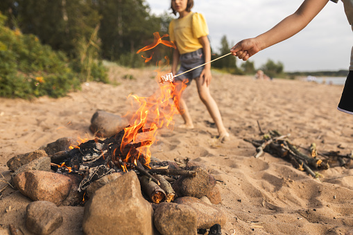 Children's hand with marshmallow on stick roasted on fire summer on beach, lifestyle