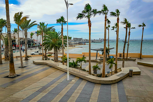 Aerial view of promenade or Malecon with palm trees, concrete benches, buildings, vehicular street, cars, beach, port and sea in background, cloudy spring day in La Paz, Baja California Sur Mexico