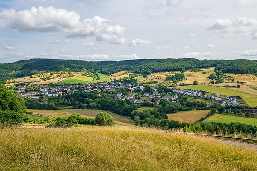 Panoramic landscape of German countryside with small village and farmland, green leafy trees covering a hill in background, sunny day in Eiffel Kreis, Bitburg-Prüm district in Rijnland-Palts, Germany