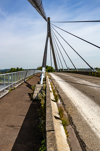 Lanaye cable-stayed bridge, vehicular road and pedestrian or bicycle path, tower, pylon, huge turnbuckles and cables, misty blue sky in background, wild plants in pavement, sunny day in Vise, Belgium
