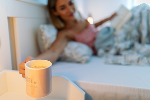 Young woman lying on bed, reading a book and drinking coffee or tea. She is putting coffee mug on a nightstand.