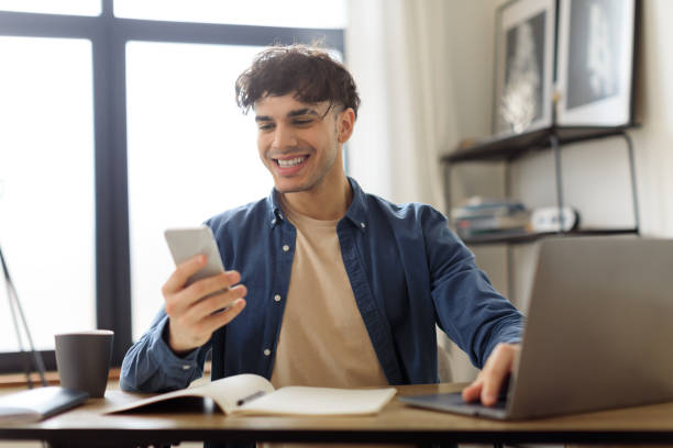istock Cheerful Guy Using Smartphone Texting Sitting At Laptop In Office 1769051508