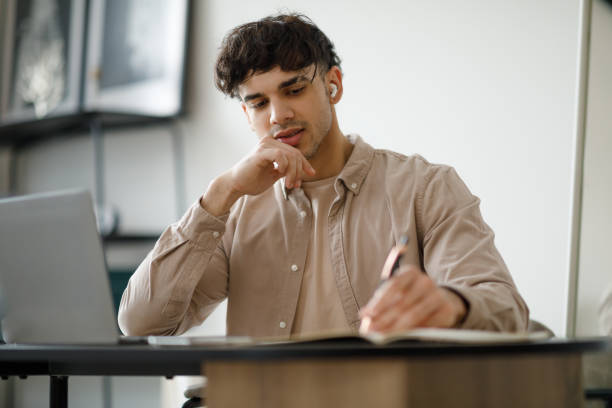istock Man Taking Notes Sitting At Laptop Wearing Earbuds At Home 1769051518