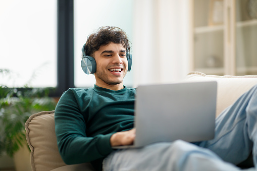 E-Learning Offer. Cheerful Arabic Guy Typing And Websurfing On Laptop Wearing Wireless Earphones Sitting On Sofa At Home. Student Having Fun And Learning Online Using Computer
