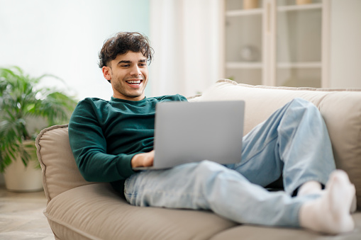 Middle Eastern guy works online at laptop with comfort resting on sofa at home. Student typing and surfing the web in modern living room, enjoying e-learning and internet fun