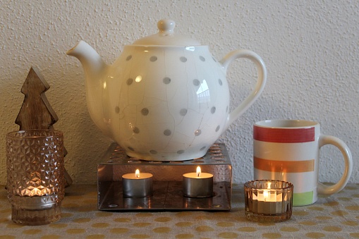 Tea pot on a pot warmer, tea cup with tealights and a wooden Christmas tree for decoration.