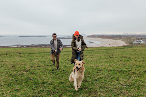 A wide angle front view of a young couple and their golden retriever dog enjoying a coastal dog walk in Newton by the Sea in the North East of England. They are wrapped up for the cold weather.