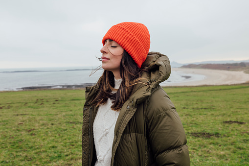 A medium close up of a young woman enjoying a coastal walk with her partner. She is having a mindful moment and closing her eyes and feeling the sea air on her face. She is in Newton by the Sea in the North East of England.