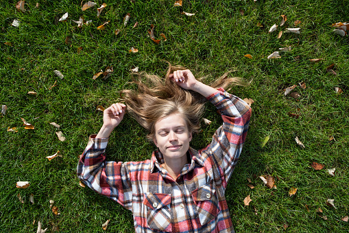 young caucasian woman in red plaid shirt smiling while relaxing on green grass with yellow autumn leaves, top view