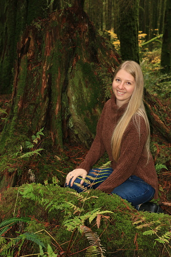 A Caucasian female model kneeling in ferns next to a tree in a public forest in Autumn. She is wearing long, blond hair, a big smile and a brown sweater.