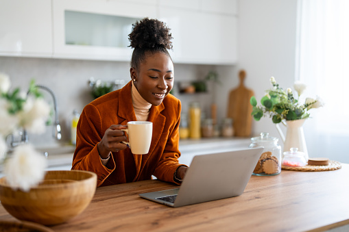 Smiling successful African woman entrepreneur working on laptop while having coffee in home office.