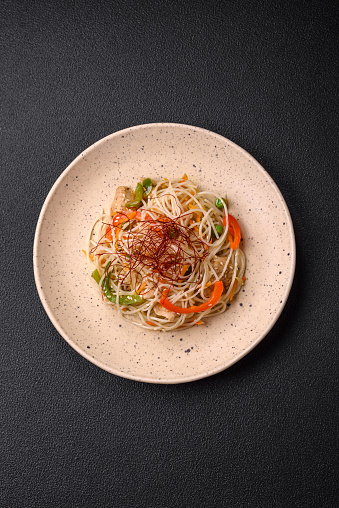Tasty dish of Asian cuisine with rice noodles, chicken, asparagus, pepper, sesame seeds and soy sauce on dark concrete background