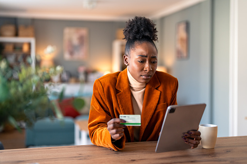 Worried young adult woman holding credit card while checking electronic bank account over digital tablet pc.