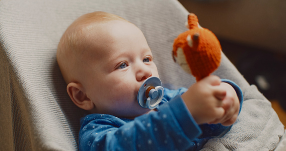 A close-up of our cute baby boy,gazing at a toy while holding a pacifier in his mouth on the bouncer at home. Pure innocence and curiosity