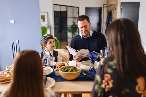 Smiling Jewish father reading Hebrew Bible during traditional Hanukkah dinner at the dining table