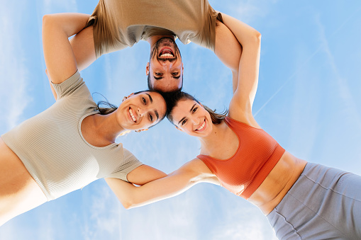 Low angle view of young group of three fitness friends in circle smiling at camera against blue sky.