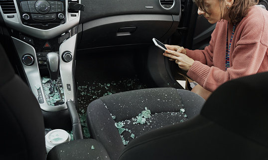 Young woman taking photos using a smart phone of the inside of her car covered in broken window glass after a break in