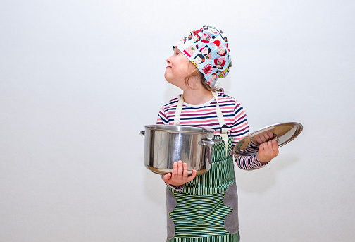 A Caucasian girl, 8 years old, dressed as a cook, holds a large saucepan in her hands and doesn’t like it. Emotional face.