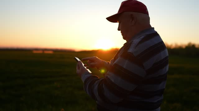 Old senior man farmer with smartphone working a in field. Senior farmer is engaged in smart farming agriculture. The tractor is running in the background.