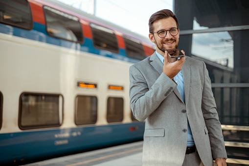 Smiling Middle Age Businessman Using a Smart Phone Voice Recognition on Line While Standing in Station Platform and Waiting for High-speed Train or Metro