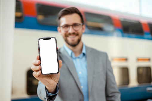 App Ad. Happy Smiling Man Passenger Holding Smartphone With White Blank Device Screen in Hand Close Up to Camera in a Train Station. Gadget With Empty Free Space for Mockup