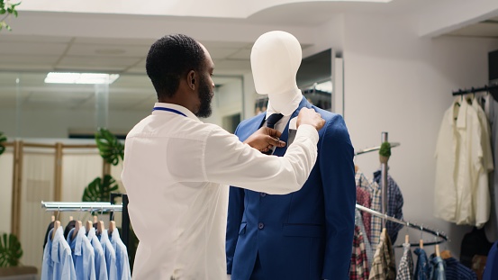 Stylish employee arranging elegant clothes on mannequins in luxurious fashion boutique. Worker adding white pocket square to high street men blazer in fancy clothing store