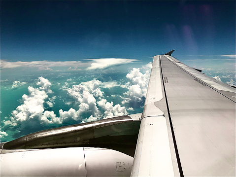Airbus A321 flying over the Atlantic Ocean. The ocean is blue and the sky has a blue with big puffy clouds. The engine is visible. The clouds cast a shadow on the water and an island is in the distance.