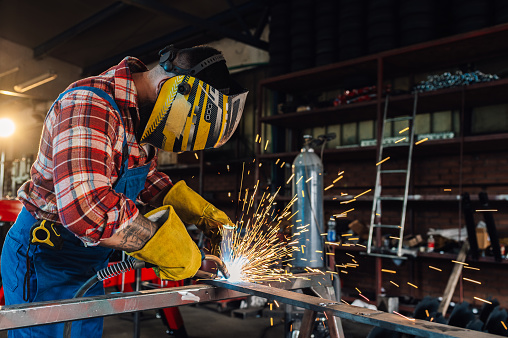 Man in a safety gear with a welding mask and yellow gloves welding a long metal pipe while sparks fly. Crafting sturdy frames for industrial machinery, ensuring safety and precision. Copy space.