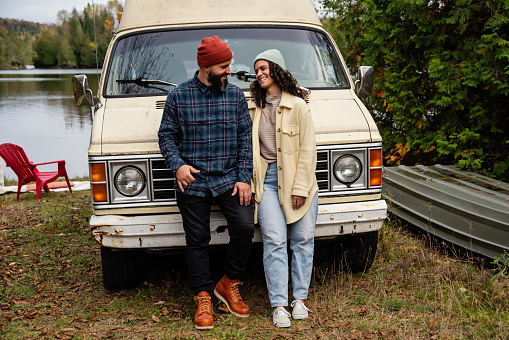 Millennial couple portrait in front of in vintage camper van. They are casually dressed with warm clothes. Lake in the background. Horizontal full length outdoors shot with copy space. This is part of a series and was taken in Quebec, Canada.