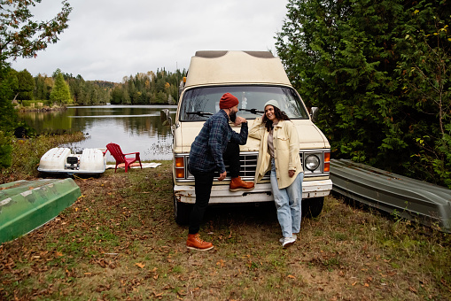 Millennial couple portrait in front of in vintage camper van. They are casually dressed with warm clothes. Lake in the background. Horizontal full length outdoors shot with copy space. This is part of a series and was taken in Quebec, Canada.