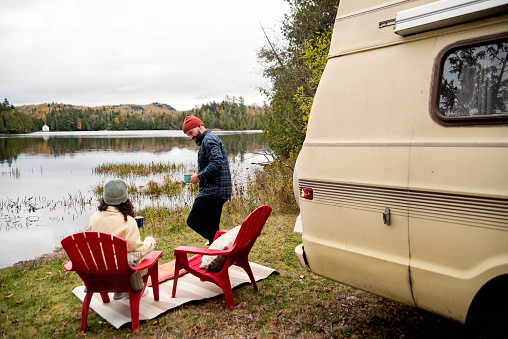 Millennial couple enjoying a week-end in vintage camper van. They are looking at the lake view with a drink. They are casually dressed with warm clothes. Horizontal full length outdoors shot with copy space. This is part of a series and was taken in Quebec, Canada.